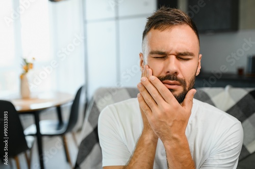 Toothache. Unhappy upset caucasian sad man sits on the couch at home, holds his hand near his cheek, grimaces, has an acute toothache, needs a consultation with a dentist doctor