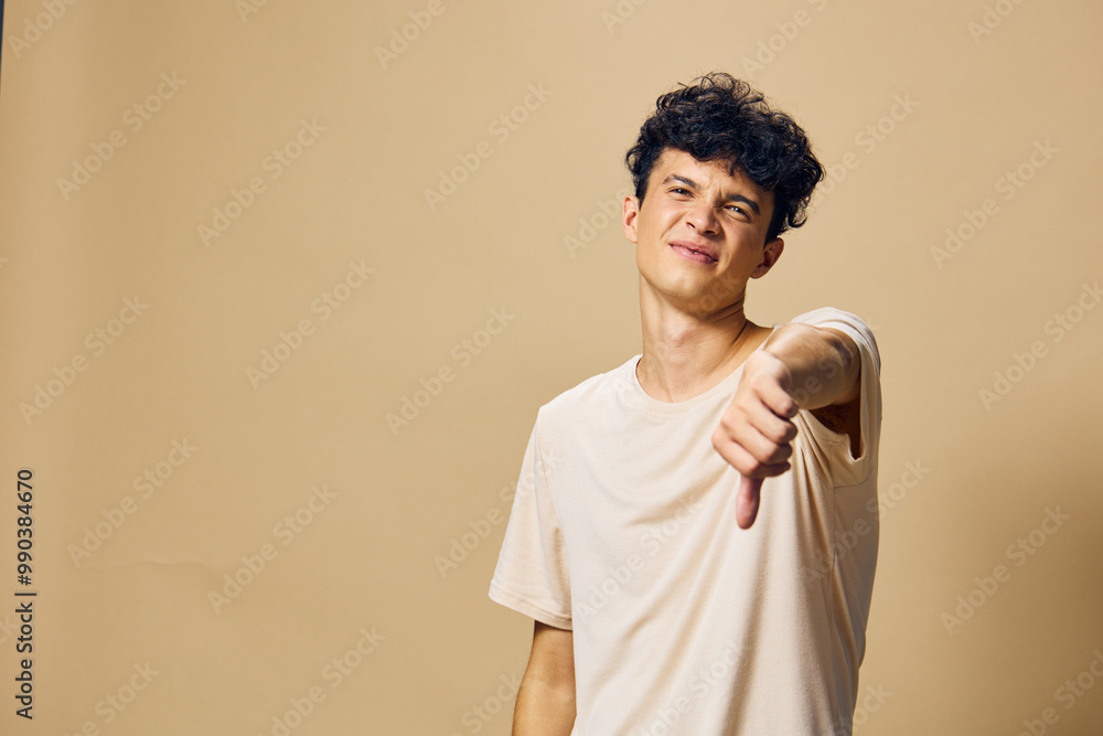 Young man showing a thumbs down gesture against a beige background, expressing disapproval and dissatisfaction with a lighthearted demeanor, making for an engaging image Young man showing a thumbs down gesture against a beige background, expressing disapproval and dissatisfaction with a lighthearted demeanor, making for an engaging image