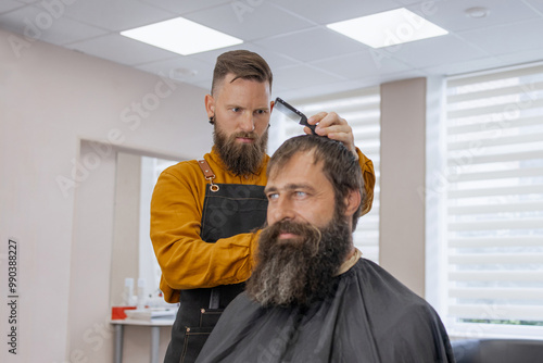 Stylish handsome barber in orange clothes and apron doing haircut to man and trimming beard in barber shop