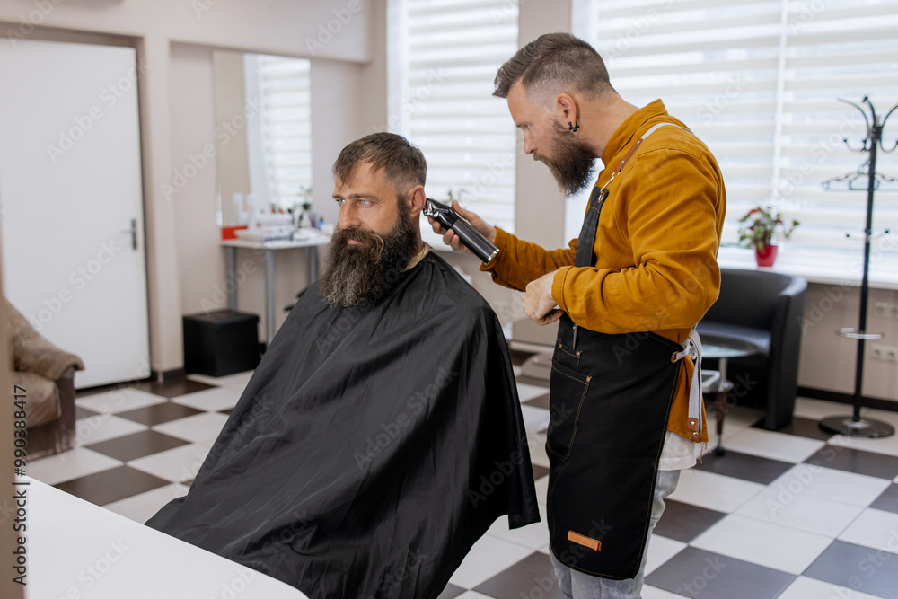 Stylish handsome barber in orange clothes and apron doing haircut to man and trimming beard in barber shop Stylish handsome barber in orange clothes and apron doing haircut to man and trimming beard in barber shop