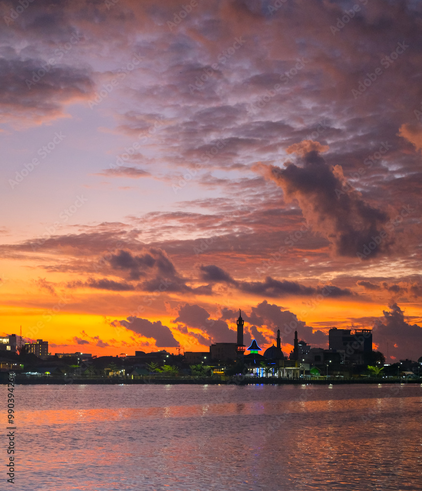 Sunset over Kapuas River in Pontianak, illuminating the sky with orange, pink, and purple hues, reflecting on calm waters. City skyline and mosque silhouette enhance the evening scenery.