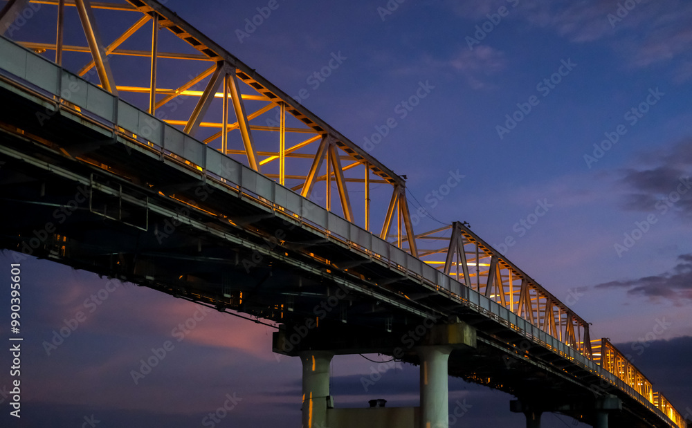 The Kapuas River Bridge in Pontianak glows beautifully at sunset, reflecting warm lights on the water. The scene captures the serene transition from day to night, showcasing urban charm.