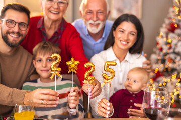 Multi-generation family holding balloons shaped as numbers 2025 while having Christmas dinner