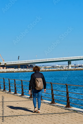 Person walking along a waterfront promenade in Cádiz on a clear day, with a view of a bridge, calm water, and blue sky in the background photo