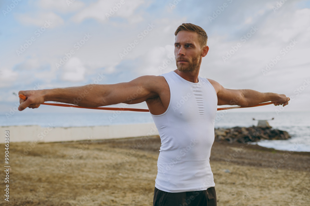 Young strong sporty athletic fit sportsman man he wearing sports clothes use elastic rubber band for hands warm up training at sunrise sun dawn over sea beach outdoor on seaside in summer day morning.