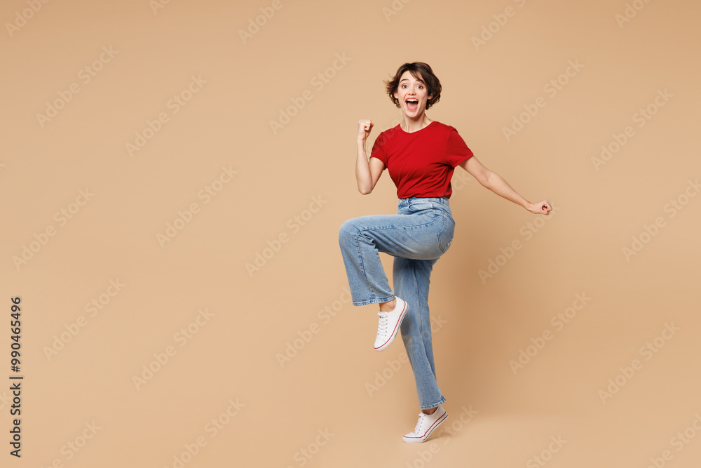 Full body young happy woman wear red t-shirt casual clothes doing winner gesture celebrate clenching fists say yes isolated on plain pastel light beige background studio portrait. Lifestyle concept. Full body young happy woman wear red t-shirt casual clothes doing winner gesture celebrate clenching fists say yes isolated on plain pastel light beige background studio portrait. Lifestyle concept.