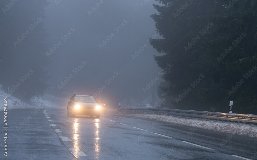A car with headlights on is driving up the mountain road in dark and foggy winter conditions. A car with headlights on is driving up the mountain road in dark and foggy winter conditions.