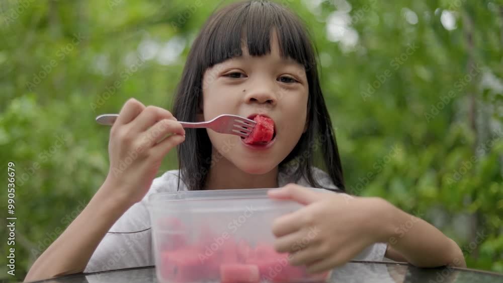 A cute Asian girl is using a fork to eat pieces of red watermelon in the front garden of her house as a fruit break during the day. in different poses, delicious and happy to eat her favorite fruit. A cute Asian girl is using a fork to eat pieces of red watermelon in the front garden of her house as a fruit break during the day. in different poses, delicious and happy to eat her favorite fruit.