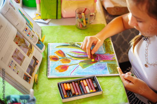 A little girl happily draws in a sketchbook at a table littered with colored pencils and pastels in a bright, sunny cheerful setting. Drawing of flowers