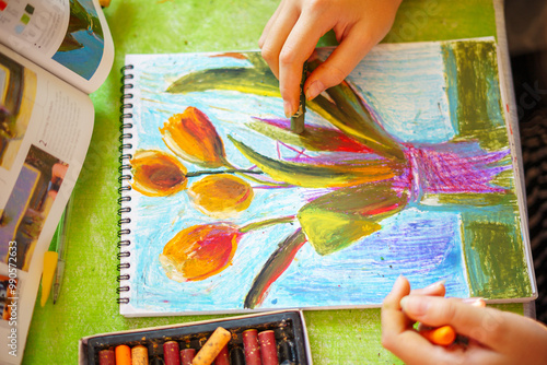 A little girl happily draws in a sketchbook at a table littered with colored pencils and pastels in a bright, sunny cheerful setting. Drawing of flowers