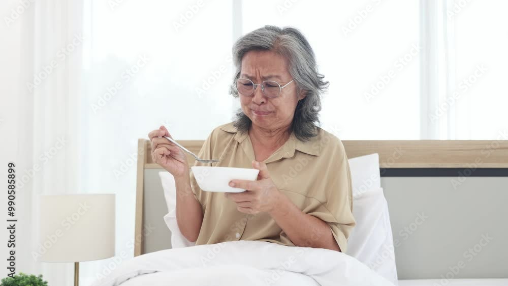 Elderly asian woman holds bowl breakfast with rejected expression on her face. She looks at the bowl food and is bored with eating the food in the bowl that is bland lacks flavor and is monotonous. Elderly asian woman holds bowl breakfast with rejected expression on her face. She looks at the bowl food and is bored with eating the food in the bowl that is bland lacks flavor and is monotonous.