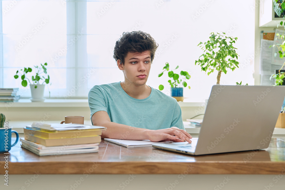 Fototapeta premium Young male college student sitting at desk at home typing on laptop