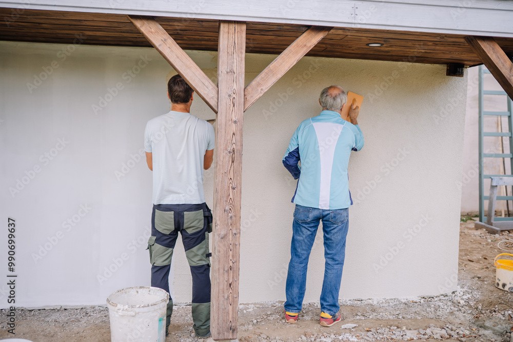 Two men plastering an exterior wall, seen from the back, working together on a home renovation project outdoors. Two men plastering an exterior wall, seen from the back, working together on a home renovation project outdoors.