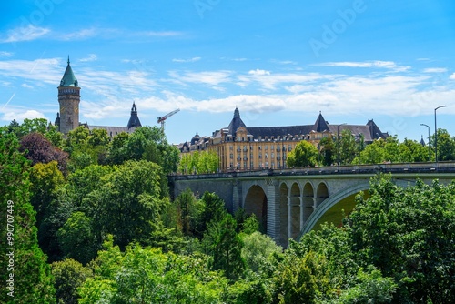 A beautiful summer day in Luxembourg City, featuring the iconic Notre-Dame Cathedral and Adolphe Bridge. The sky is clear blue, with lush green trees and a mix of old and new architecture. photo