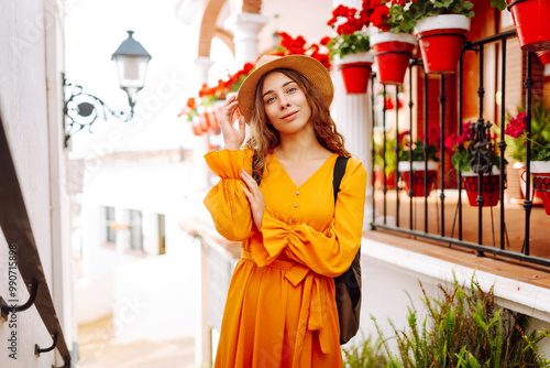 A young traveler woman in a vibrant orange dress and hat enjoys a sunny stroll through a charming white-washed village with flower pots. Concept of lifestyle, vacation, fashion, travel #990715898