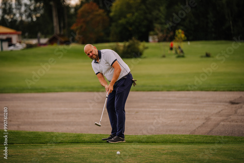 A golfer prepares to putt on the green, focusing intently on the ball. The background includes a golf course and surrounding greenery.