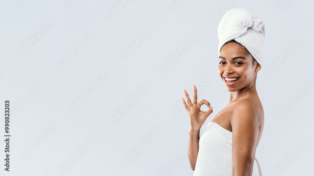 Satisfied young afro woman wrapped in towels showing okay gesture and smiling over grey background, copy space, body care products Satisfied young afro woman wrapped in towels showing okay gesture and smiling over grey background, copy space, body care products
