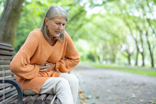 Schilderij op canvas Elderly woman sitting on park bench outside experiencing stomach discomfort