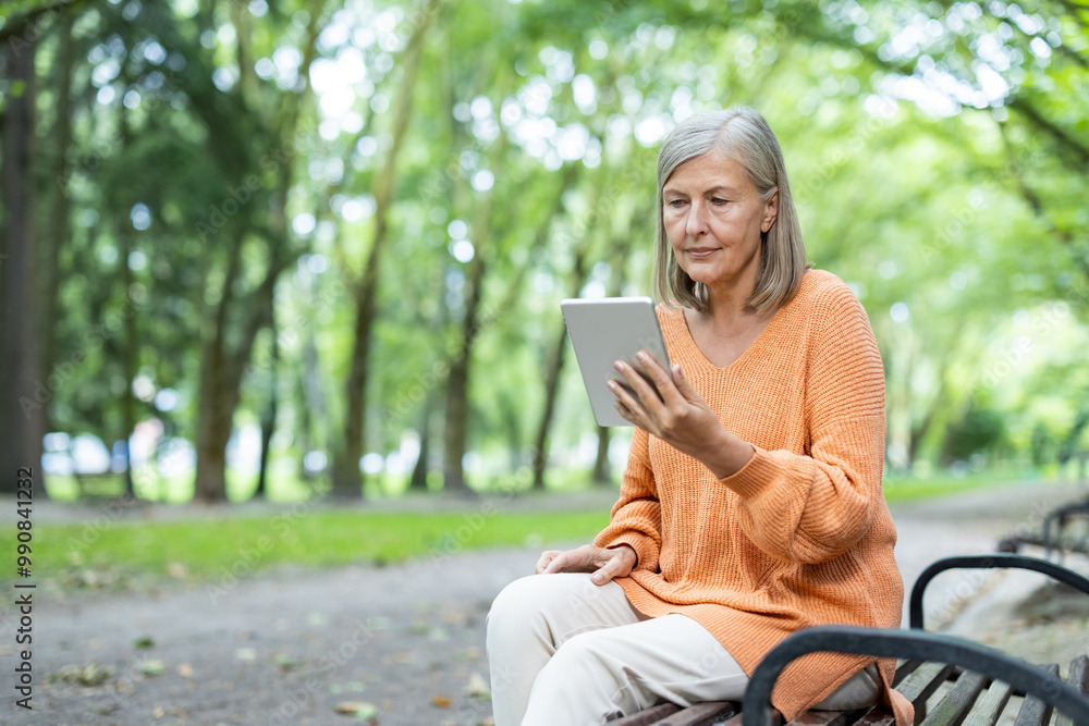 Older woman seated on park bench using tablet, engaging with digital technology. Outdoor setting emphasizes tranquil lifestyle, blending modern technology with nature. Older woman seated on park bench using tablet, engaging with digital technology. Outdoor setting emphasizes tranquil lifestyle, blending modern technology with nature.