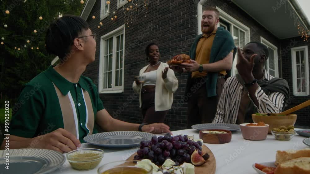 Stab shot of multiracial friends having dinner party on patio in backyard on summer evening. Two diverse male friends having conversation at table while their friend serving main dish in front of them Stab shot of multiracial friends having dinner party on patio in backyard on summer evening. Two diverse male friends having conversation at table while their friend serving main dish in front of them