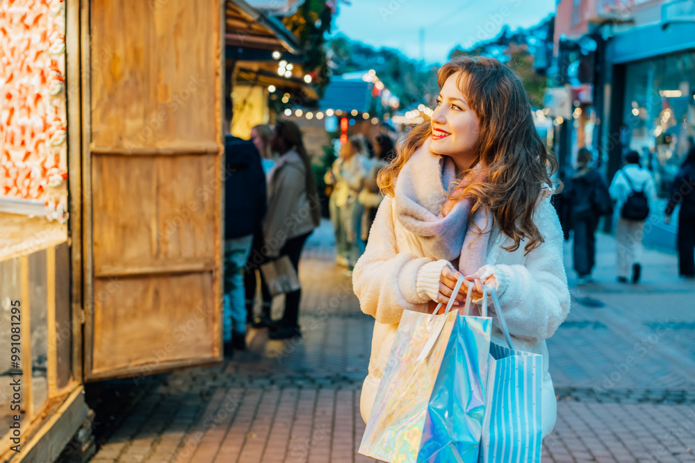 Fototapeta premium Happy woman enjoys shopping at festive Christmas market while carrying shopping paper bags, walking in street Xmas fair of European city. Selecting gifts for New Year, winter holiday sales, discounts