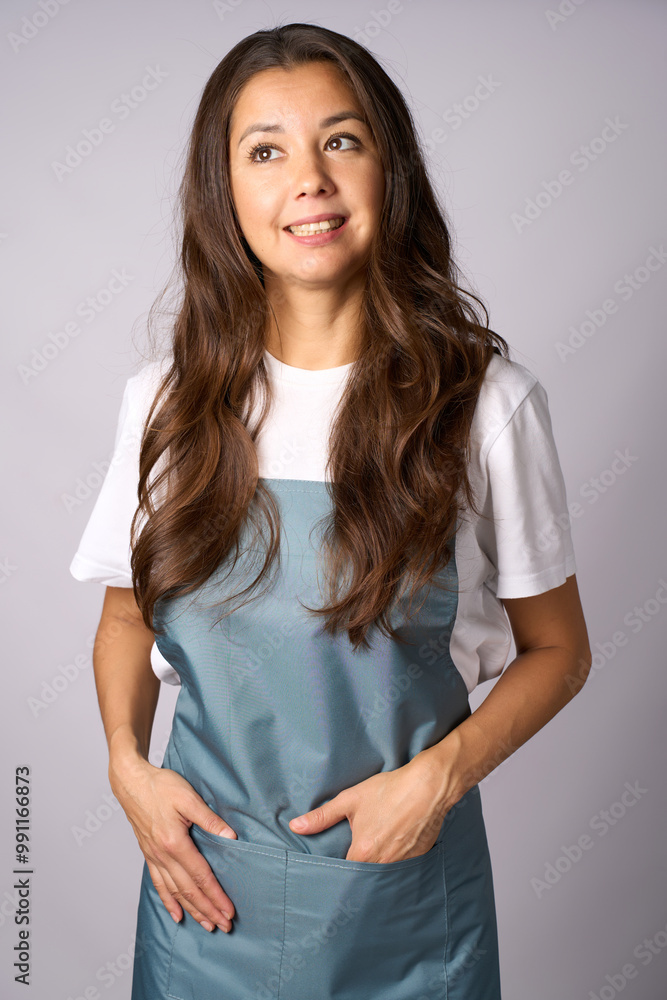 Friendly female barista in a apron standing with hands in pockets, smiling against a light gray background Friendly female barista in a apron standing with hands in pockets, smiling against a light gray background