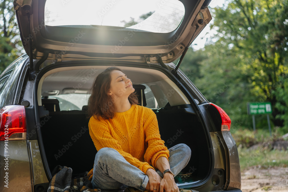 Young cheerful woman traveler sitting in open trunk of car enjoying sunset in forest