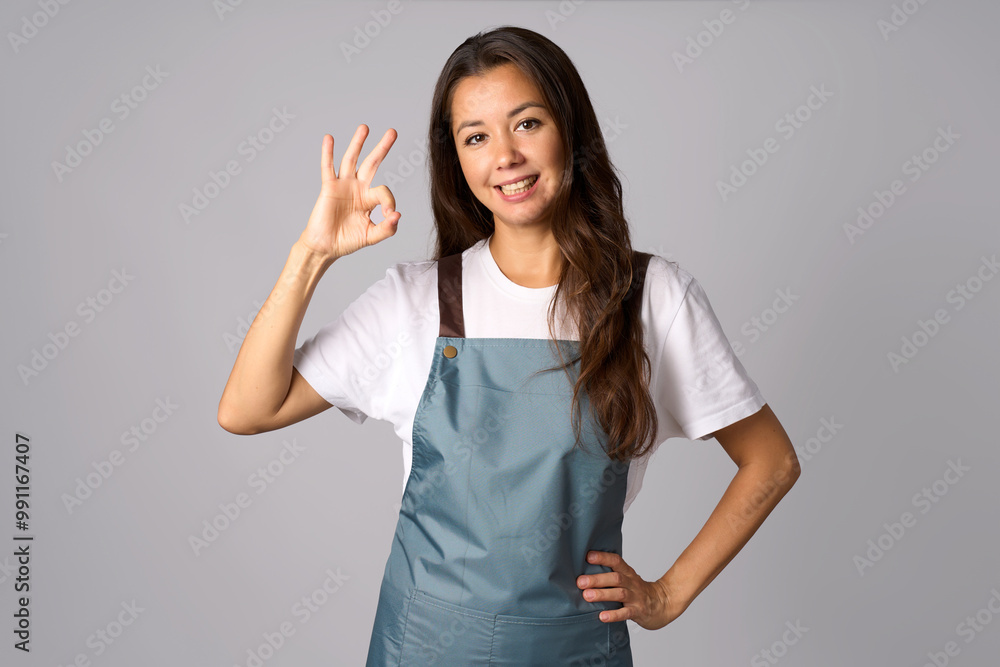 Beautiful waitress showing ok sign, wearing apron and standing against gray background Beautiful waitress showing ok sign, wearing apron and standing against gray background