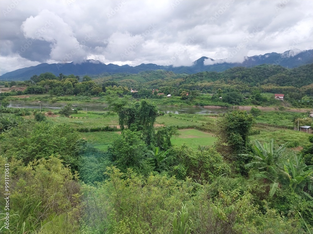 Obraz premium Scenic view of lush green valley with mountains under cloudy sky, captured in rural landscape.