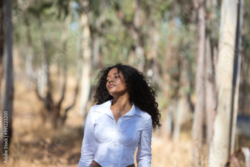 Woman, young, beautiful, Latin, brunette with curly hair dressed in a white shirt with her head tilted back and her eyes closed, she is relaxed. The woman is among the eucalyptus trees in the forest.