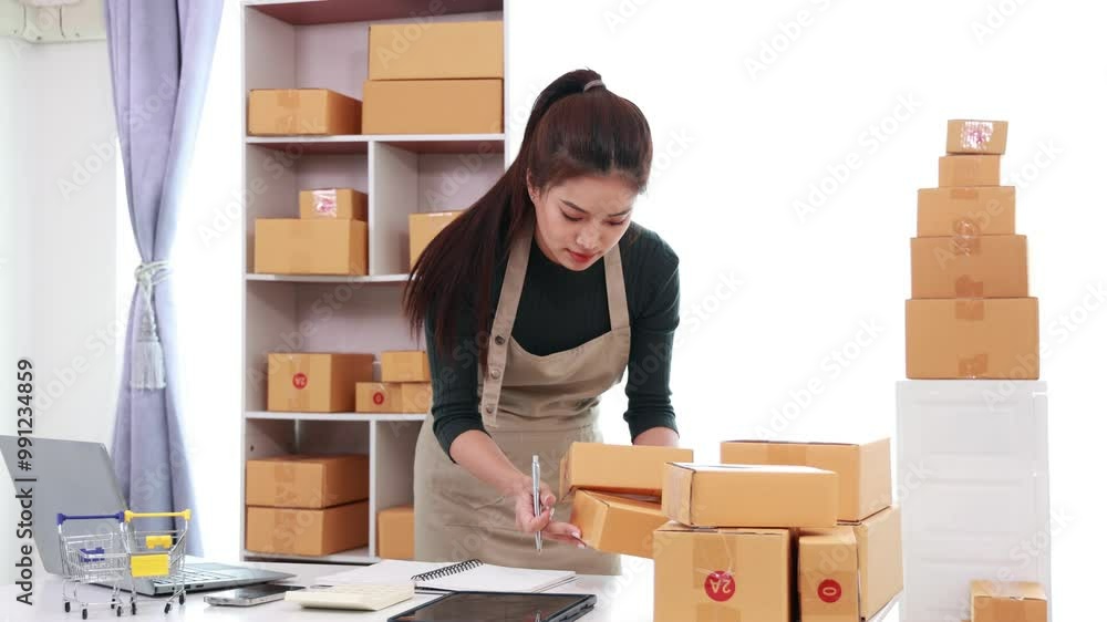 An Asian businesswoman works from home, managing her online business through her laptop and smartphone. She efficiently handles online orders, packaging, and shipping. An Asian businesswoman works from home, managing her online business through her laptop and smartphone. She efficiently handles online orders, packaging, and shipping.