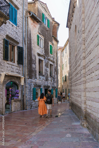 Wanderers explore the narrow, cobblestone streets of Kotor, surrounded by ancient stone buildings adorned with vibrant shutters. Sunlight bathes the historic town in a warm glow.