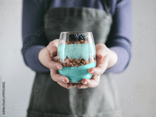 Triffle dessert in woman hands. Blue spirulina and fruit smoothie with granola, blueberry and blackberry in glass. Healthy detox food and breakfast idea and recipe. photo