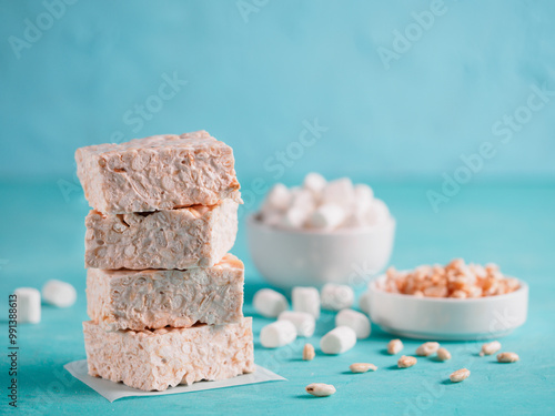 Homemade square bars of marshmallow and crispy rice with ingredients on azure blue background. Stack of American dessert with marshmallow and crispy rice. Copy space