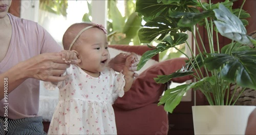 Mom teaches Asian baby girl how to walk at home. Candid shot - Mother and smiling infant laughing and enjoy each other’s company. Happy family fosters joyful and energetic child.