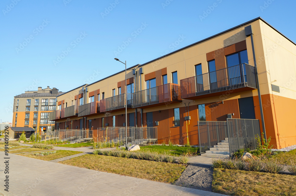 Townhouse in a suburb area. New apartment buildings in green enviroment. Street with Two-story townhouses. Modern Townhouse along sidewalk with green lawn.Pedestrian path with flowerbed shrubs. Townhouse in a suburb area. New apartment buildings in green enviroment. Street with Two-story townhouses. Modern Townhouse along sidewalk with green lawn.Pedestrian path with flowerbed shrubs.