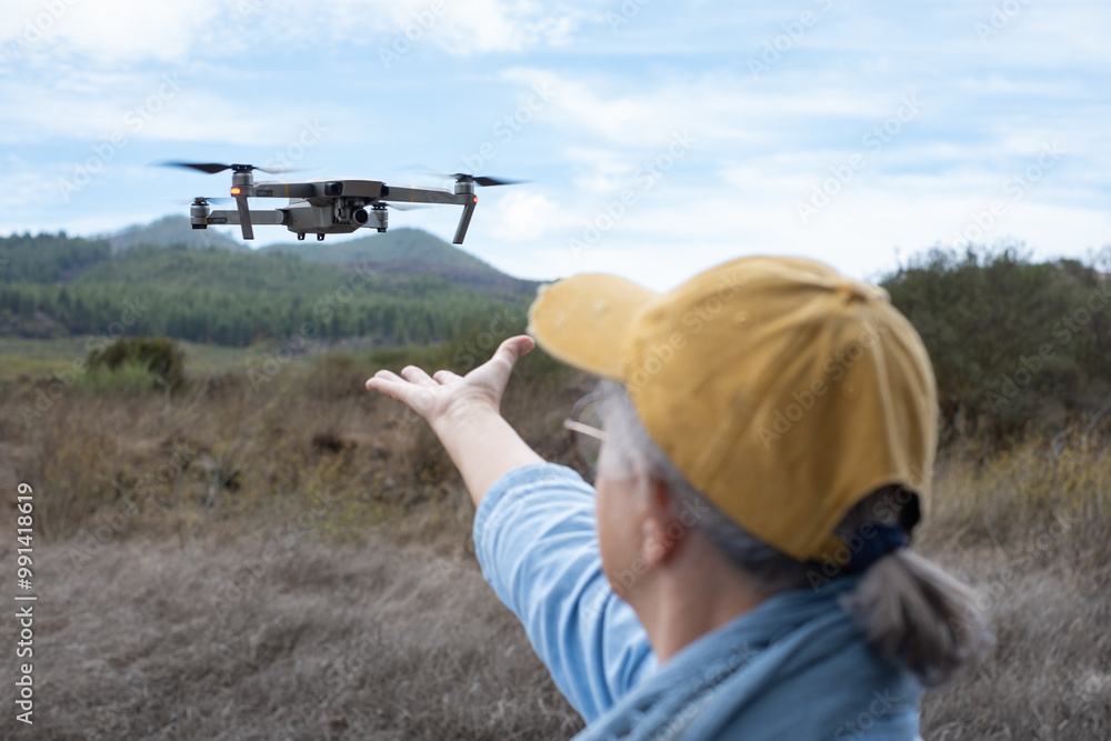 Rear view of a mature woman flying a drone using the remote control. The drone pilot releases the drone camera from the hands. Aerial nature photography and videography Rear view of a mature woman flying a drone using the remote control. The drone pilot releases the drone camera from the hands. Aerial nature photography and videography