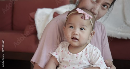 Mom and cute Asian baby girl cuddling at home. Candid shot - Mother and infant enjoy quality time together. Portrait of happy, joyful and energetic child looking curiously at the camera.