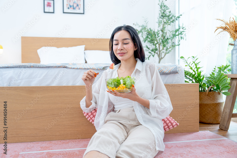 Pretty Asian woman hold bowl of vegetable salad and she express happy with eating, she also sit near bed in bedroom with relax time. Pretty Asian woman hold bowl of vegetable salad and she express happy with eating, she also sit near bed in bedroom with relax time.