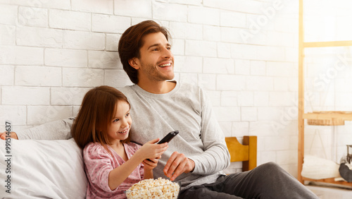 Cozy weekend at home. Cute little girl watching TV with her young dad, eating popcorn, copy space