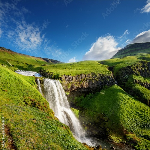 A breathtaking view of a lush hillside showcasing a cascading waterfall under a bright blue sky sprinkled with light, fluffy clouds