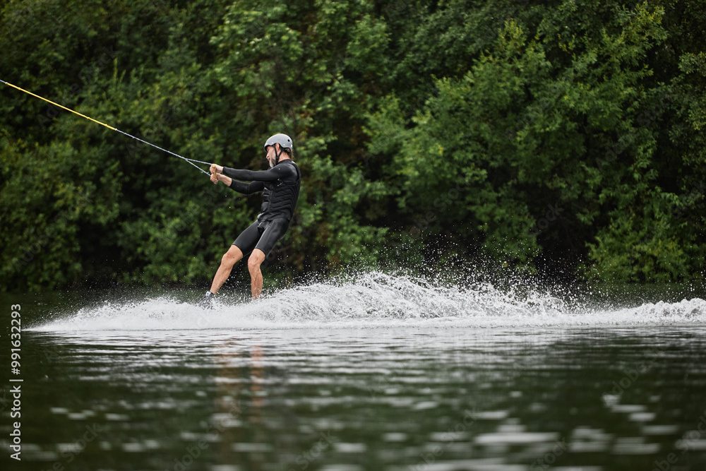 Wide angle side view of active mature man wakeboarding on lake with blurred green forest background sending wave trail copy space