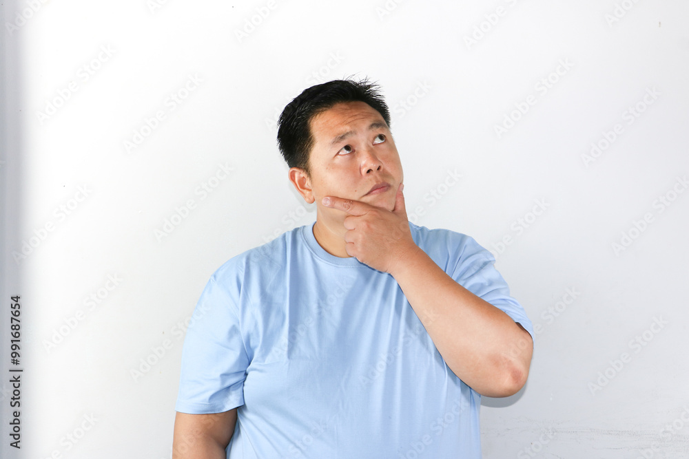 Thoughtful Asian man wearing blue t-shirt holding his chin and thinking with serious expression consider something.