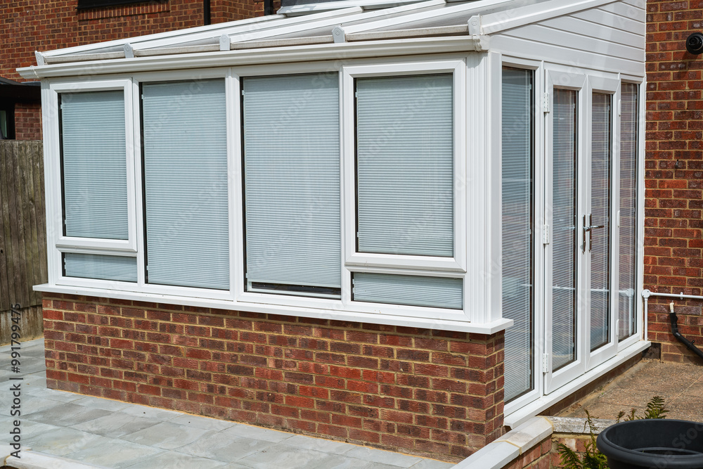 White lean to conservatory with blinds on all windows and doors, seen from the outside on a sunny day. White lean to conservatory with blinds on all windows and doors, seen from the outside on a sunny day.