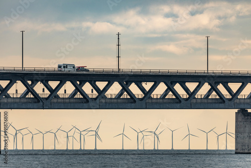A caravan crosses an elevated bridge, next to a car with a surfboard on the roof. Below, the sea is home to a wind farm, with rows of turbines standing out on the horizon.
