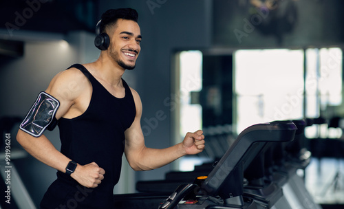 Handsome Young Muscular Arab Man In Wireless Headphones Jogging On Treadmill At Gym, Sporty Motivated Middle Eastern Guy Listening Music While Running, Enjoying Making Sports, Copy Space