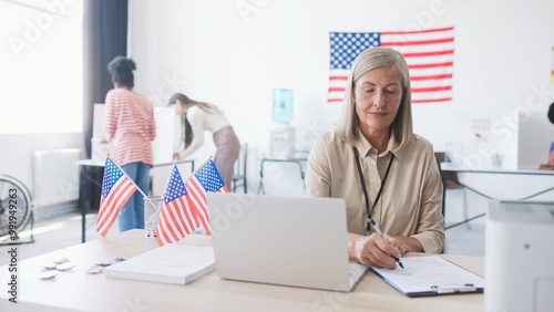 Beautiful American woman sitting in front of portative laptop in polling station office. Joyfully smiling while proudly looking at camera. People voting for Presidential candidates in background. photo