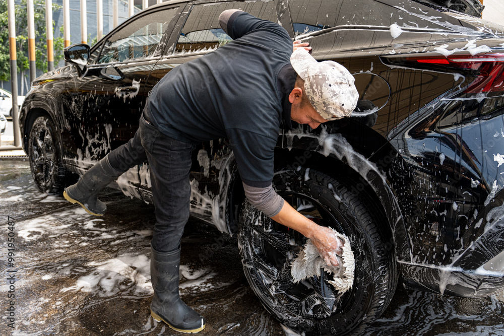 Obraz premium Latin man washing a black car with foam soap and a sponge.