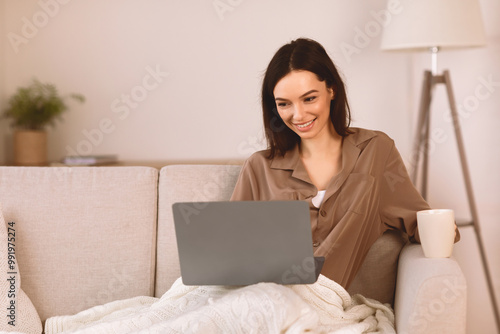 Cozy Lifestyle. Smiling woman wrapped in warm blanket lying on sofa with cup of coffee, surfing internet on her laptop