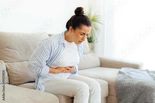 Stomach pain, problems with indigestion, menstruation. Sad arabian or indian woman sitting on a sofa, suffering stomach ache, constipation, cramp, nausea or bloated, holds his stomach with his hands